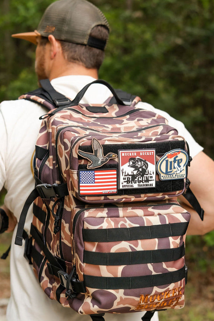A man stands outdoors facing away, wearing the Mucker Bucket Duck It Limited Edition Tac-Pac - 25L tactical backpack decorated with patches like an American flag, duck, and vintage-style logos.