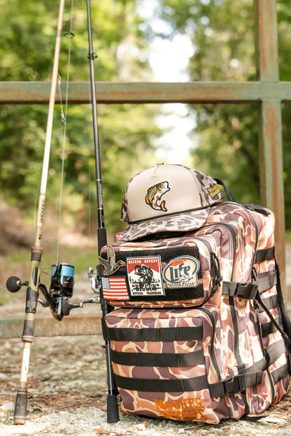 A Mucker Bucket Duck It Limited Edition Tac-Pac - 25L sits on a gravel path outdoors with two fishing rods, a fishing hat, and trees plus a metal fence in the background.