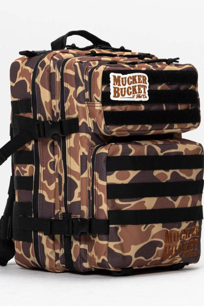 The Duck It Limited Edition Tac-Pac - 25L by Mucker Bucket is a camouflage-patterned hunting backpack with multiple compartments, black straps, and a Mucker Bucket patch on the top front pocket, shown against a plain white background.