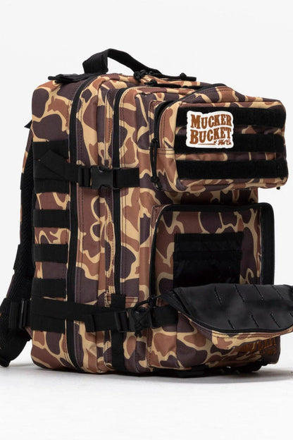The Mucker Bucket Duck It Limited Edition Tac-Pac - 25L is a camouflage-patterned backpack with multiple compartments, black straps, and a branded patch on the top front pocket. One lower pocket is open to show the interior, set against a plain white background.