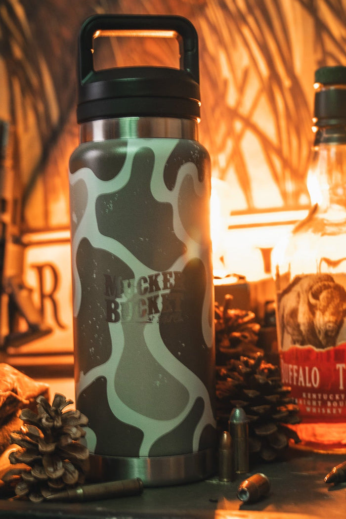 The Mucker Bucket Duck Jug - Duck Camo Water Bottle (26oz) sits amid pinecones, Buffalo Trace whiskey, bullets, and a jacket on a rustic surface. This leak-proof stainless steel bottle is perfect for every adventure.