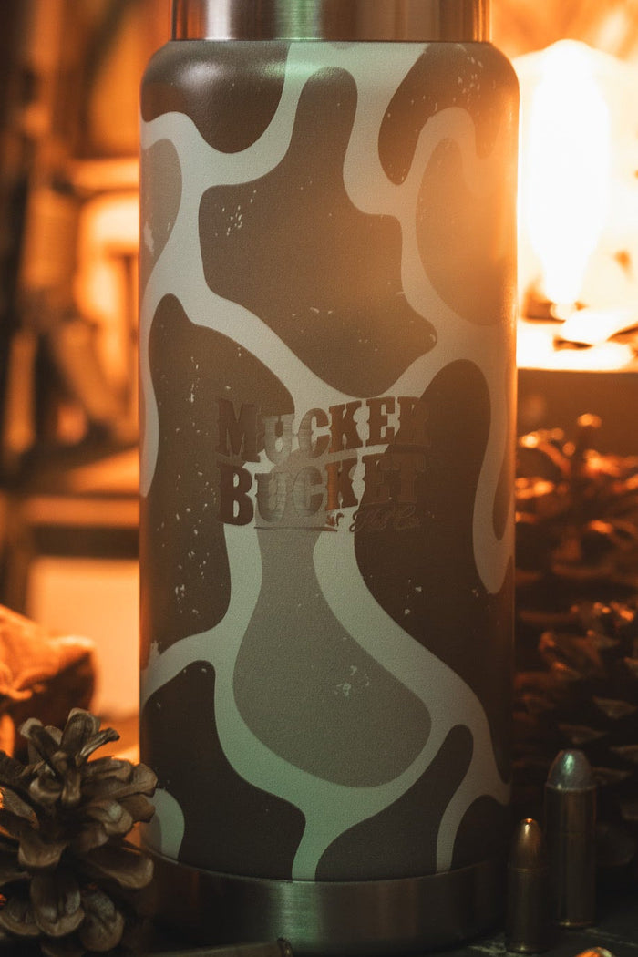 A close-up of the 26oz Duck Jug - Duck Camo Water Bottle by Mucker Bucket sits on a table with pinecones, a metal bullet, and a bottle in warm, soft lighting.