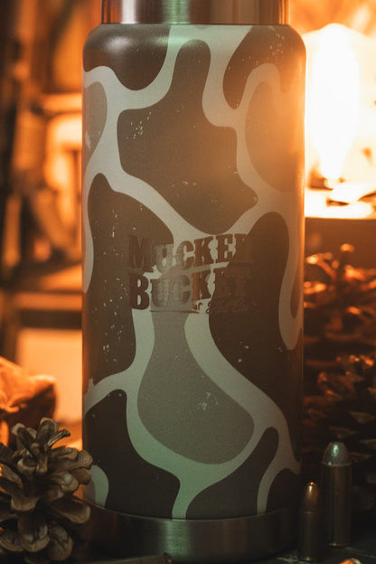 A close-up of the 26oz Duck Jug - Duck Camo Water Bottle by Mucker Bucket sits on a table with pinecones, a metal bullet, and a bottle in warm, soft lighting.