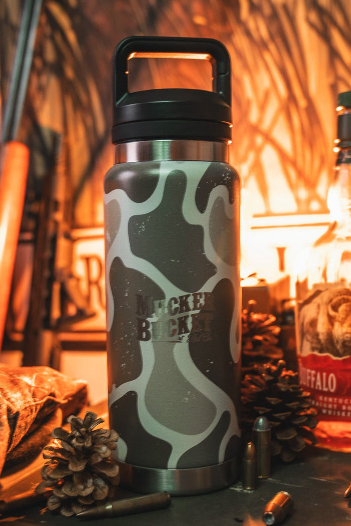 A Mucker Bucket Duck Jug - Duck Camo Water Bottle (26oz) sits on a table with pinecones, bullets, and Buffalo Trace bourbon, all set against a rustic outdoor backdrop.