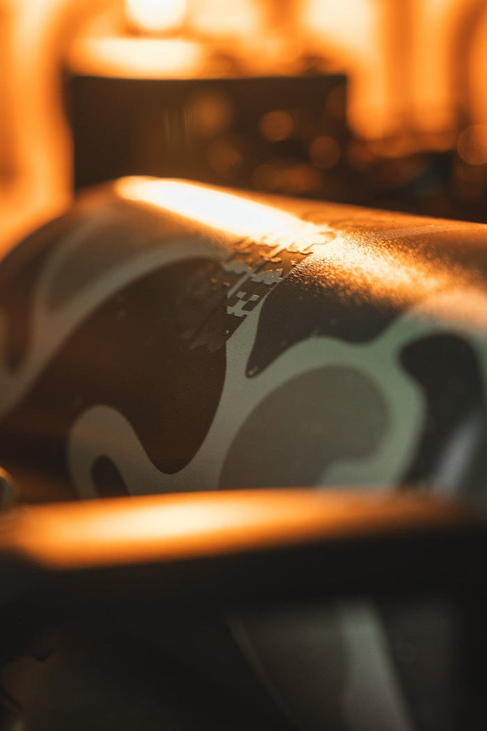 A close-up of the Mucker Bucket Duck Jug - Duck Camo Water Bottle (26oz) highlights its camouflage stainless steel body and leak-proof design, set against a blurred background with warm lighting.