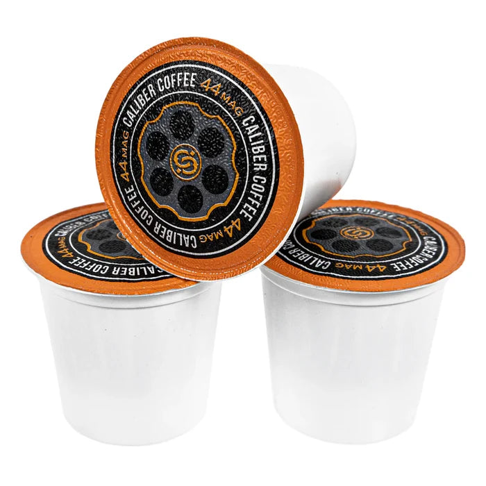 Three white coffee pods with black and orange lids are stacked together, featuring a bullet chamber design and the text 44 MAG CALIBER COFFEE around the edge. Named Espresso Roast .44 Mag Single-Serve Shells by Caliber Coffee Company, they offer specialty grade, artisan roasted precision.