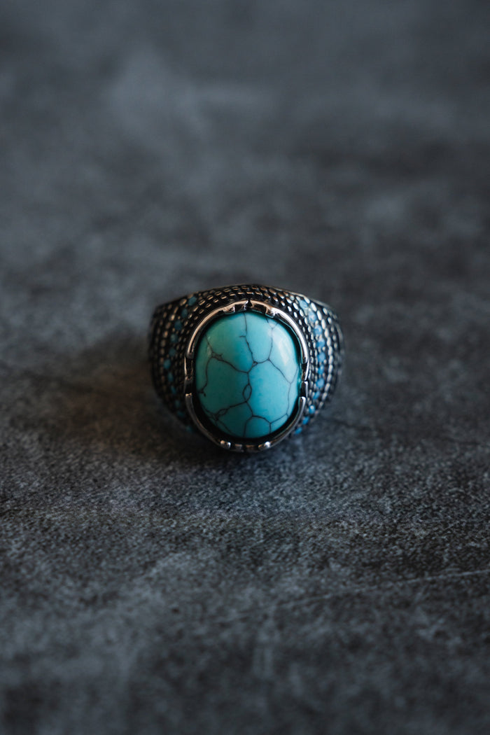 Discover Relentless Betrayals Eye of the Storm Ring, a handcrafted piece featuring a large turquoise gemstone with black veining on a dark textured background, complemented beautifully by an ornate silver band design.