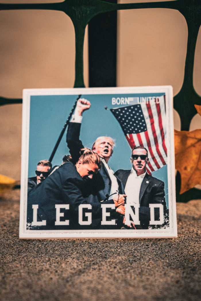 A yellow leaf lies near a card featuring the Forever Legend - Trump PVC Patch by Born United Private Label, displaying an image of a man with an American flag and bodyguards. The word LEGEND is printed on it, against a backdrop of a metal fence.