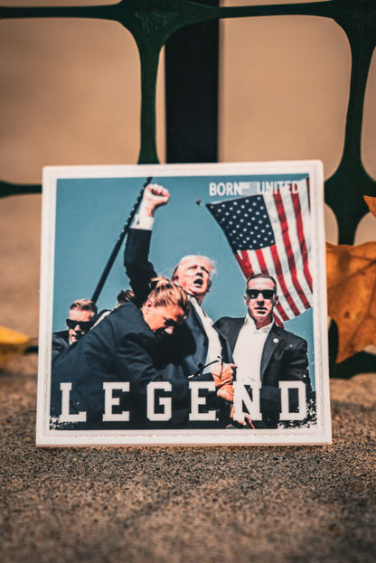 A yellow leaf lies near a card featuring the Forever Legend - Trump PVC Patch by Born United Private Label, displaying an image of a man with an American flag and bodyguards. The word LEGEND is printed on it, against a backdrop of a metal fence.