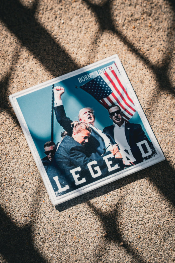 A square ground picture shows cheering people with one holding the American flag, marked LEGEND at the bottom. Shadows of a grid fall over it. This resembles the Forever Legend - Trump PVC Patch by Born United Private Label.