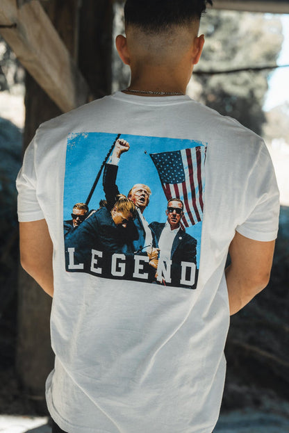 A person models the Forever Legend - Trump t-shirt by Born United Private Label, showing off its premium white fabric with a people-and-flag graphic and the text Forever Legend. The serene outdoor backdrop includes wooden structures and greenery.