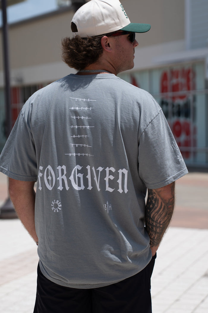 A man wearing the 13th Forgiven Premium Tee and black shorts stands outdoors on a sunny day in a shopping area, facing away from the camera. Store signs are visible, highlighting that forgiveness is possible for anyone.