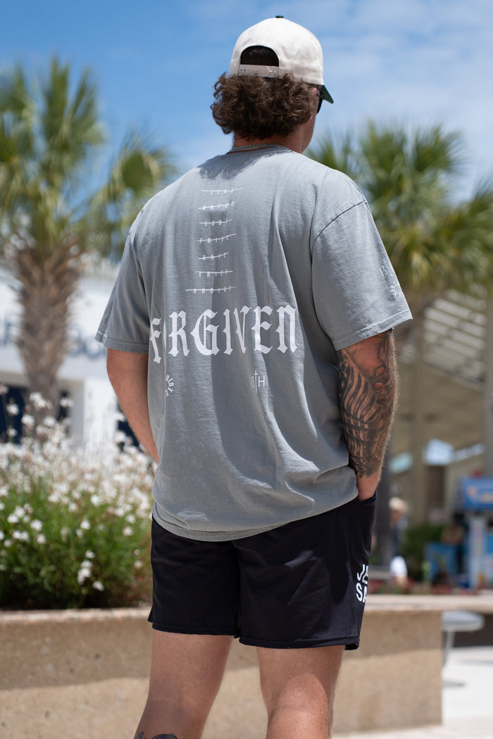 A man stands outdoors with his back to the camera, wearing a gray Forgiven Premium Tee by 13th, black shorts, a white cap, and revealing tattoos. Palm trees and buildings are visible in the background.