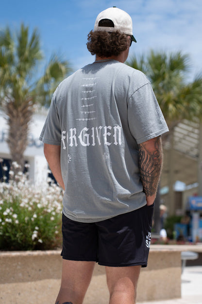 A man stands outdoors with his back to the camera, wearing a gray Forgiven Premium Tee by 13th, black shorts, a white cap, and revealing tattoos. Palm trees and buildings are visible in the background.