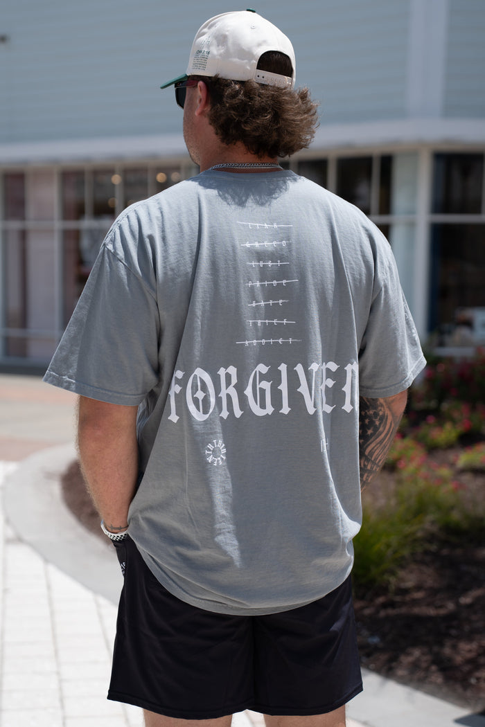 A person stands outdoors facing away, wearing the gray Forgiven Premium Tee by 13th with arrow designs, black shorts, a white cap, and sunglasses—a powerful symbol of grace and transformation from past labels.