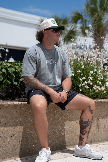 A man in the 13th Forgiven Premium Tee, black shorts, cap, and white sneakers sits on a low concrete wall near flowers. He has arm and leg tattoos and looks off to the side outdoors on a sunny day.