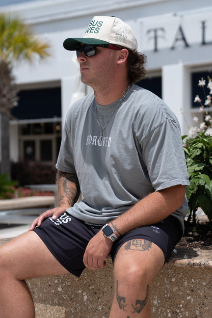 A man in a gray 13th Forgiven Premium Tee, black shorts, white sneakers, and a trucker hat sits relaxed on a concrete bench near green plants and a white building, his tattoos visible as he subtly showcases his forgiven past.