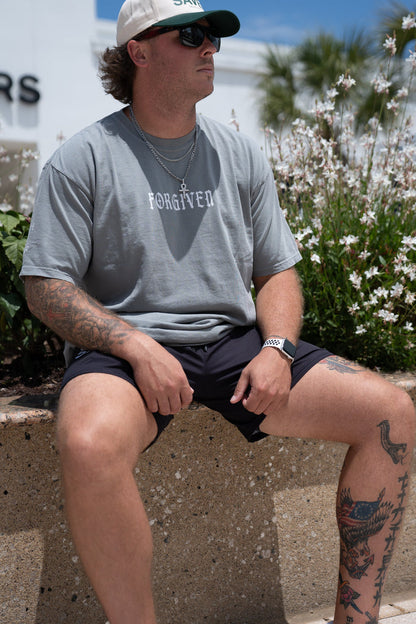 A man in a 13th Forgiven Premium Tee and JESUS SAVES cap sits outdoors on a low wall. He wears sunglasses, shorts, and white sneakers, showing tattoos like "ex-addict" on his arms and legs. Flowers and greenery are in the background.