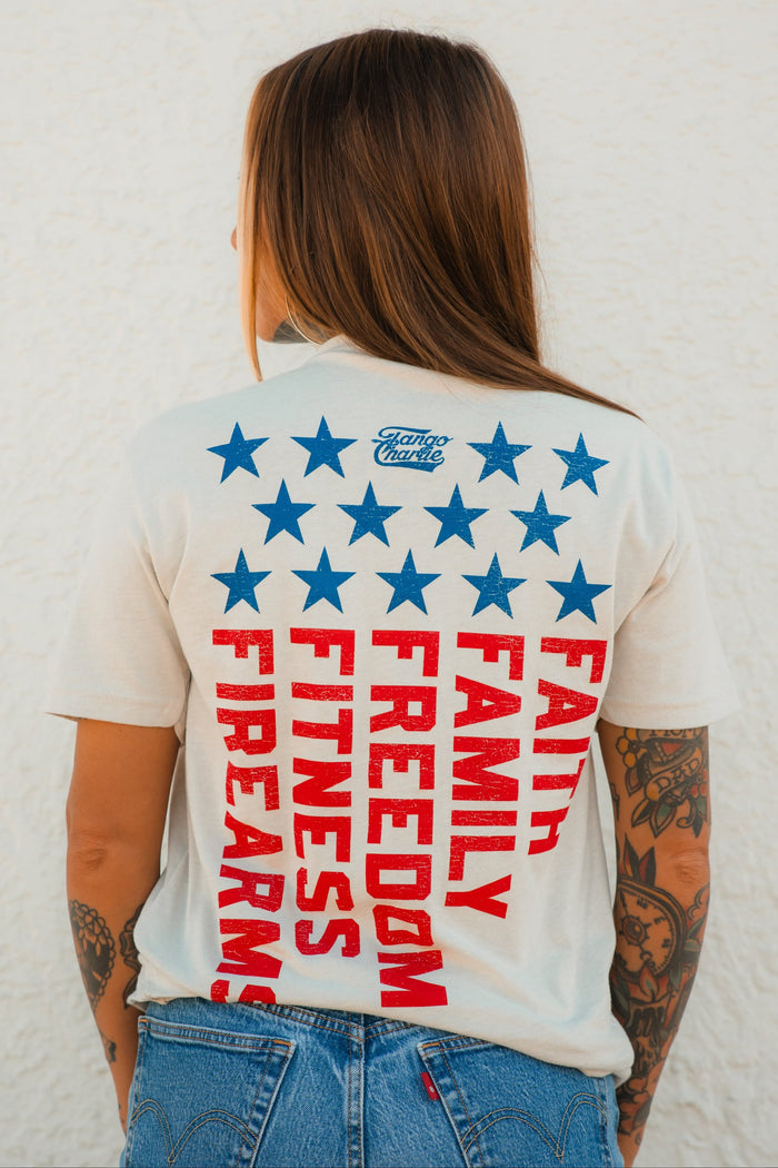 A person with long brown hair wears the Tango Charlie Apparel Foundational Five - Tee, a white shirt with blue stars and bold red “Faith Family Freedom” text. Visible arm tattoos and blue jeans complete the look, seen from behind.