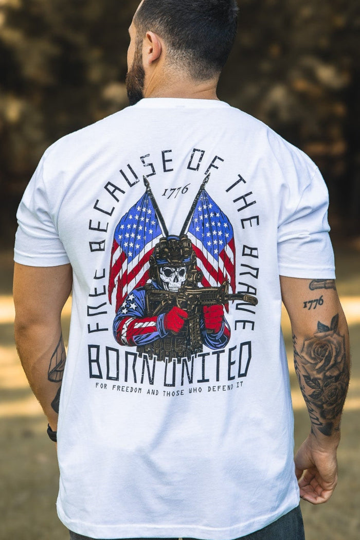 A tattooed man outdoors, back to the camera, wears a white Freedom Defender T-shirt from Born United Private Label featuring a skeleton with boxing gloves, American flags, and the words "Free Because of the Brave – Born United 1776.