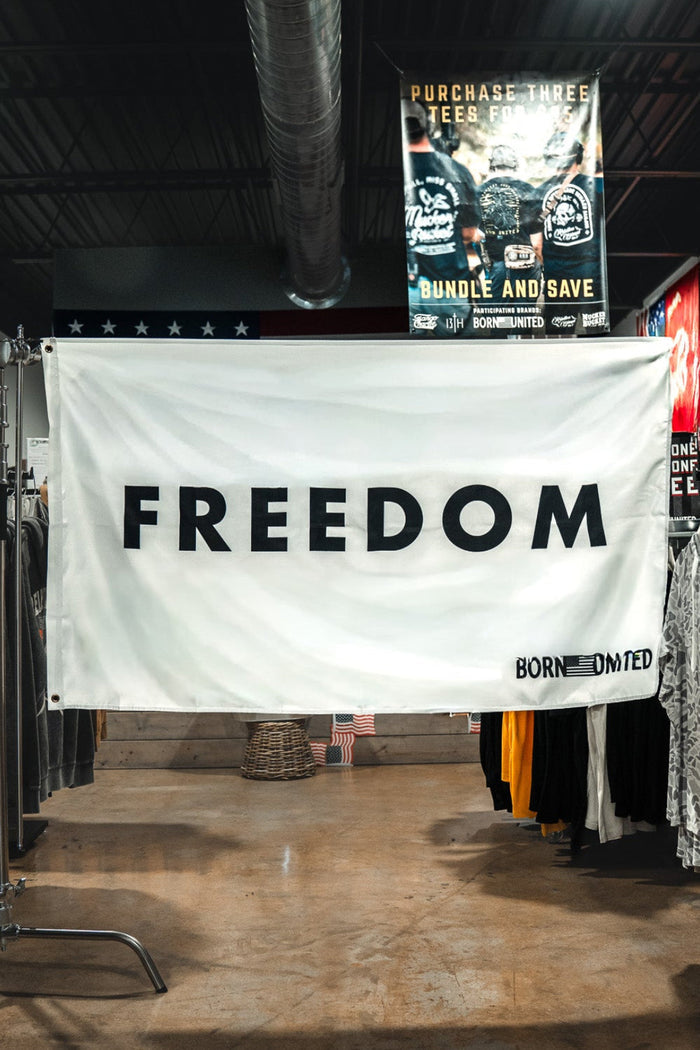 A large white Freedom Flag by Born United Private Label with bold black "FREEDOM" lettering hangs between clothing racks in a store, reflecting patriotic values. Various shirts and posters can be seen in the background.