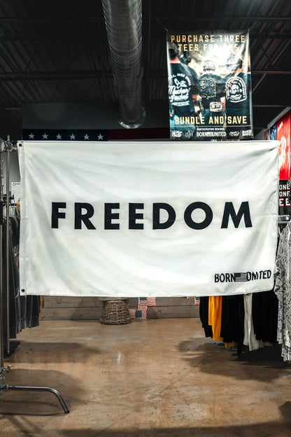 A large white Freedom Flag by Born United Private Label with bold black "FREEDOM" lettering hangs between clothing racks in a store, reflecting patriotic values. Various shirts and posters can be seen in the background.