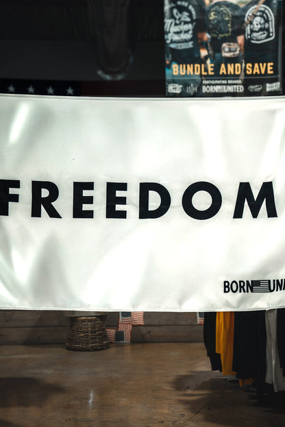 A white Freedom Flag by Born United Private Label hangs indoors with bold black "FREEDOM" and an American flag corner featuring "BORN UNITED," embodying patriotism. Clothing racks and a store sign appear in the background.