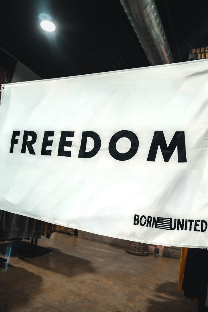 The Freedom Flag by Born United Private Label features bold black "FREEDOM," "BORN UNITED," and an American flag graphic on a white background, honoring patriotic values and American heritage in the bottom right corner.