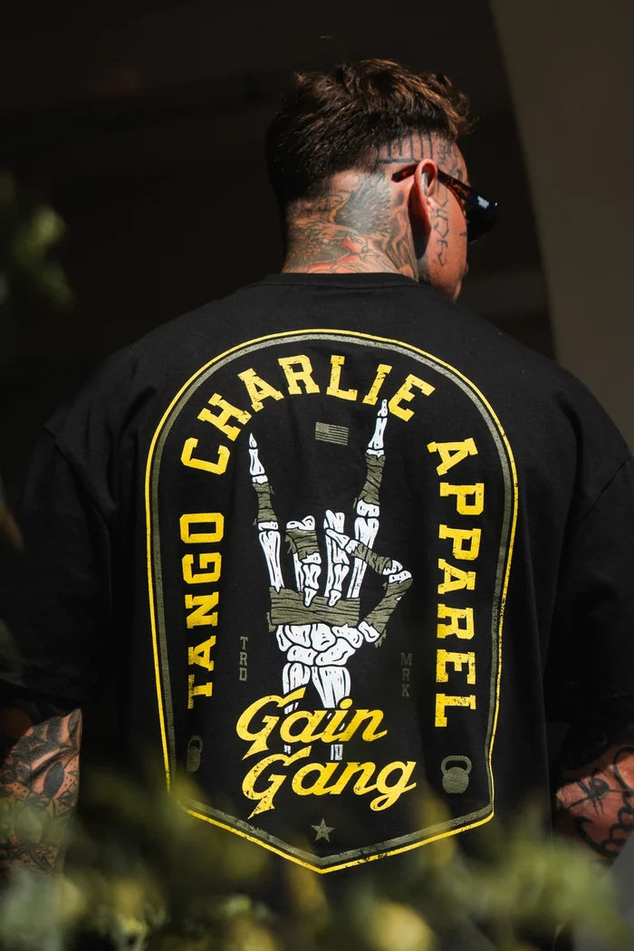 A tattooed individual with short hair and sunglasses models the Tango Charlie Apparel Gain Gang - Premium Oversized Tee, a black unisex heavyweight t-shirt with a bold yellow-outlined skeleton hand and prominent Gain Gang branding.