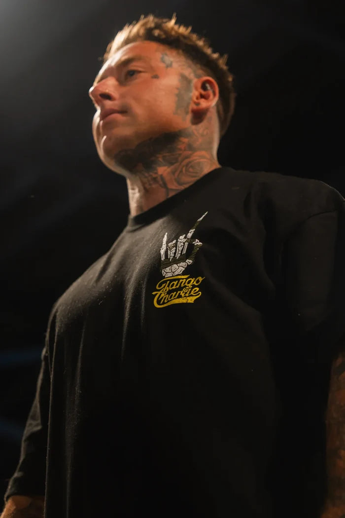 A person with face and neck tattoos wears the Gain Gang - Premium Oversized Tee by Tango Charlie Apparel, featuring a bold yellow and white graphic, standing under dramatic lighting.