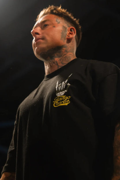 A person with face and neck tattoos wears the Gain Gang - Premium Oversized Tee by Tango Charlie Apparel, featuring a bold yellow and white graphic, standing under dramatic lighting.