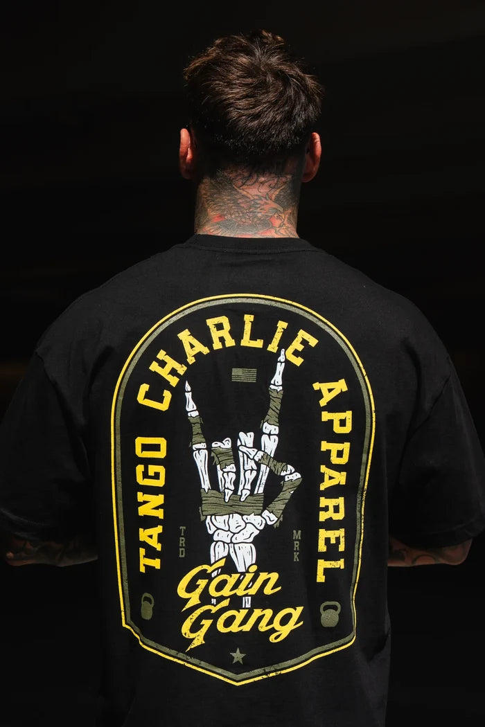 A person with neck tattoos faces away, wearing the Gain Gang - Premium Oversized Tee by Tango Charlie Apparel, featuring a large, colorful back graphic with a hand sign and bold lettering.