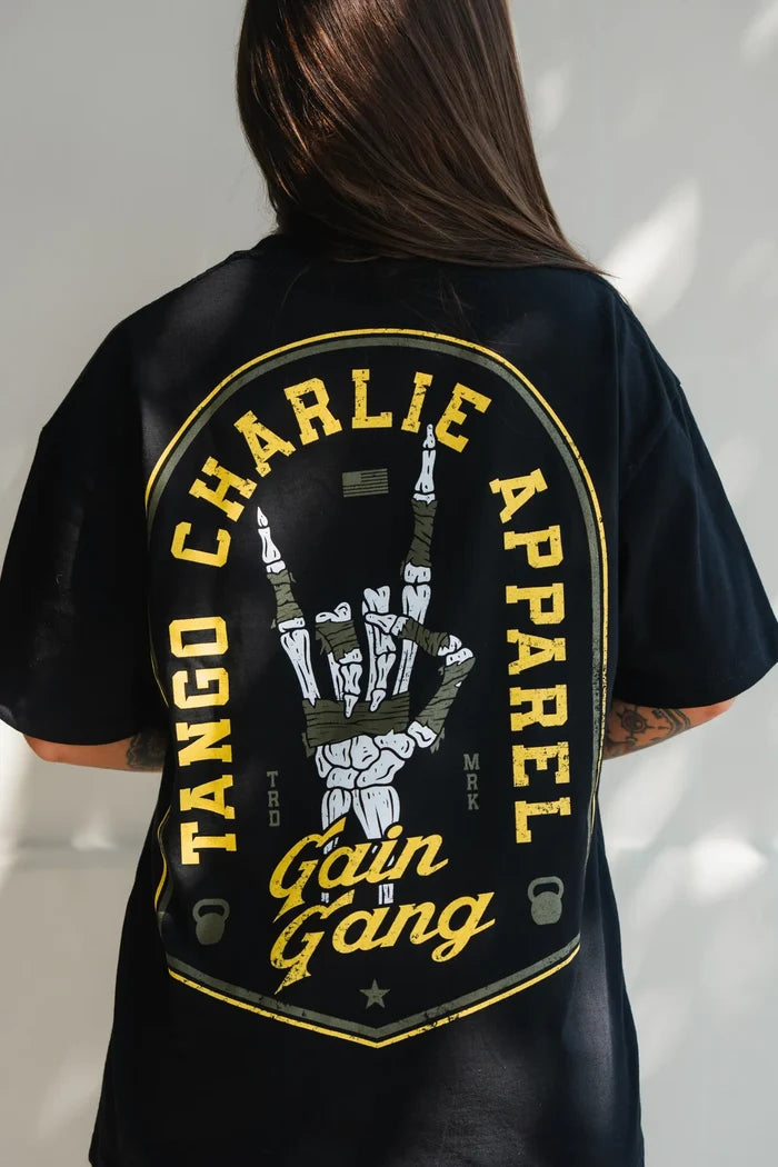 Someone wears the Gain Gang - Premium Oversized Tee by Tango Charlie Apparel, a black oversized shirt with bold yellow lettering and a skeletal hand graphic on the back, featuring standout design and premium details.