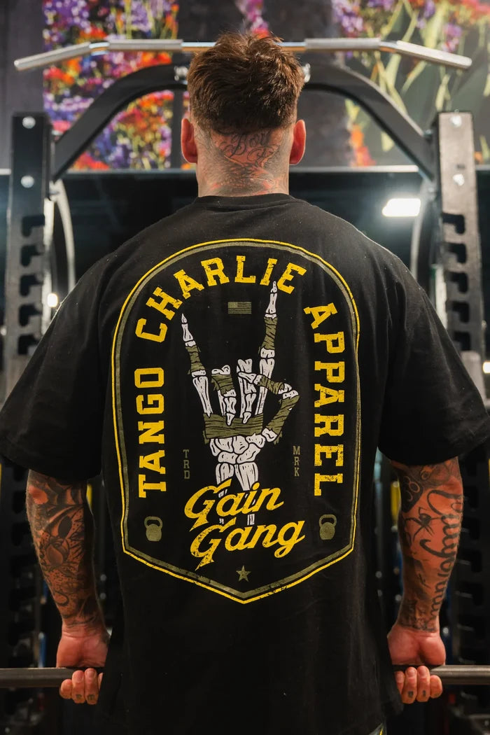 A tattooed man stands in a gym facing away, wearing the Tango Charlie Apparel Gain Gang - Premium Oversized Tee with a skeleton hand and yellow text. Gym equipment and colorful wall art are visible in the background.