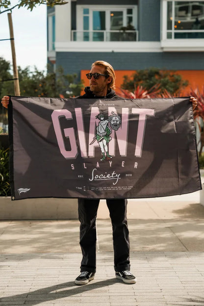 A man in sunglasses holds the Barnabas Clothing Giant Slayer Banner, which reads "GIANT SLAYER Society" with an armored character slaying a giant. Urban buildings and plants appear in the background.