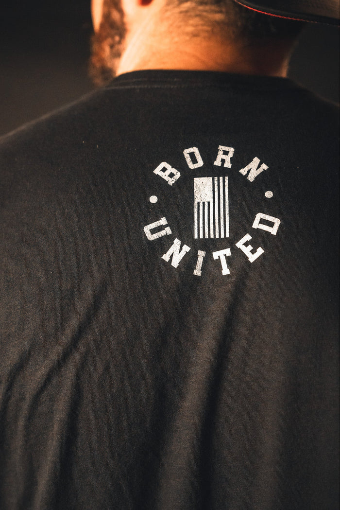 A person proudly stands wearing a black God Over Government T-shirt by Born United Private Label, featuring an American flag. The design embodies the spirit of good conduct and honors the values upheld by governing authorities.