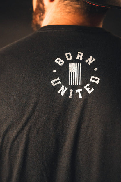 A person proudly stands wearing a black God Over Government T-shirt by Born United Private Label, featuring an American flag. The design embodies the spirit of good conduct and honors the values upheld by governing authorities.