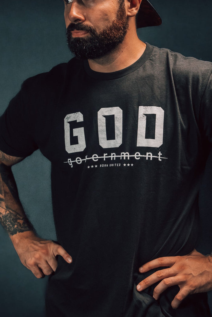 A bearded man, gazing to the side against a dark background, stands confidently with hands on hips. Hes wearing a reversed baseball cap and Born United Private Labels God Over Government t-shirt, which features the word government crossed out below GOD, challenging authority with his defiant posture.