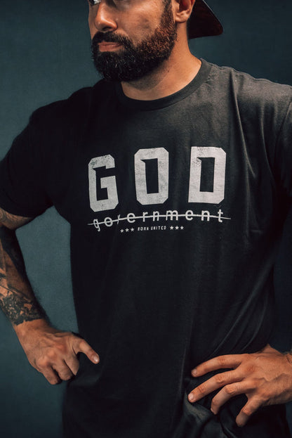 A bearded man, gazing to the side against a dark background, stands confidently with hands on hips. Hes wearing a reversed baseball cap and Born United Private Labels God Over Government t-shirt, which features the word government crossed out below GOD, challenging authority with his defiant posture.