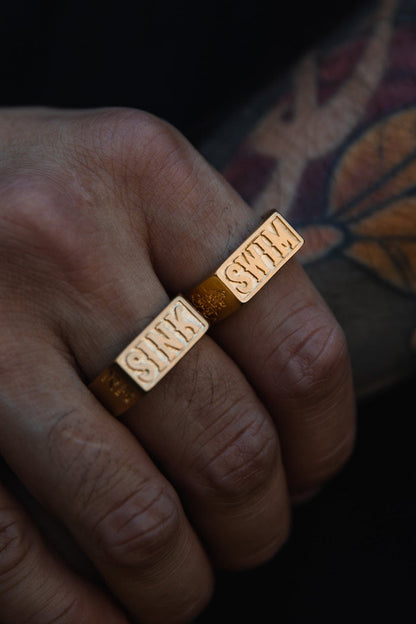 A hand wearing two Relentless Betrayals Gold Sink or Swim Rings, each engraved with SINK, rests side by side. The bold rings are complemented by a colorful tattoo on the skin, showcasing a unique blend of art and style.