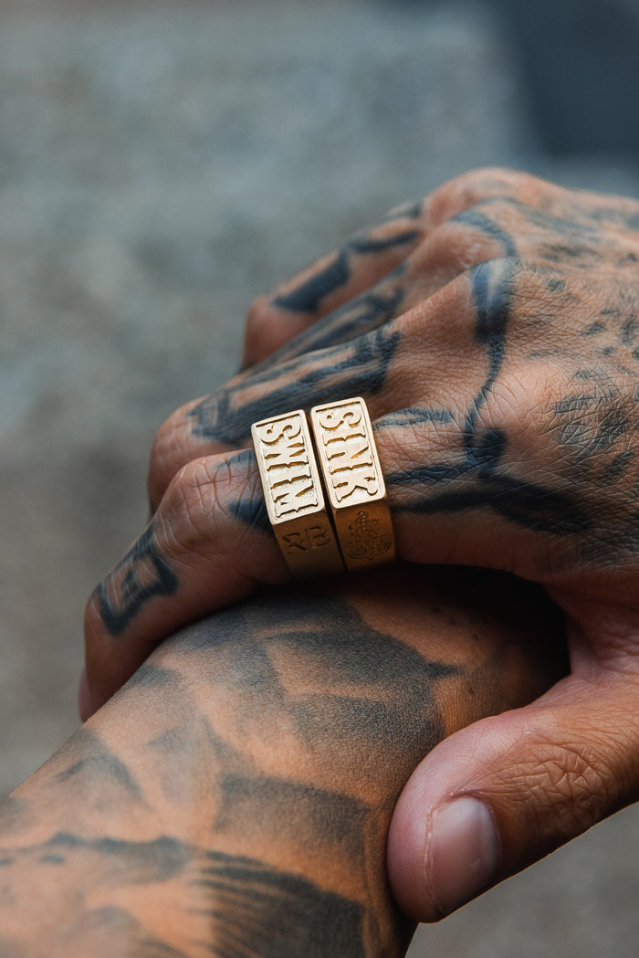 Close-up of tattooed hands, one holding the other. The hand displays two thick handcrafted Gold Sink or Swim Rings from Relentless Betrayal, with intricate SKTS engravings and tattoos featuring complex patterns against a softly blurred background.