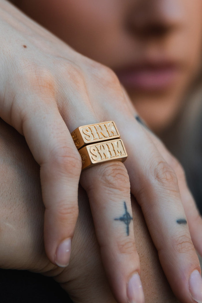 A close-up shows a hand resting on another, wearing two Relentless Betrayals handcrafted Gold Sink or Swim Rings. The fingers feature simple tattoos, like a cross on the middle finger, with a softly blurred background enhancing this intimate scene.