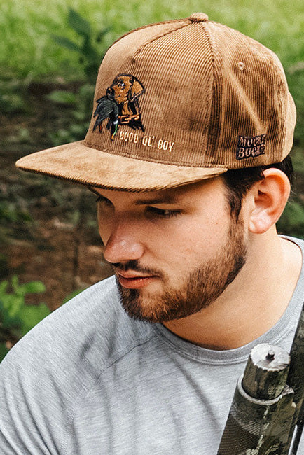 Man wearing a brown cap with a logo, holding a bow in a forest setting