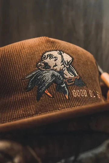 The Mucker Bucket Good Ol Boy Corduroy Bucket is a brown corduroy hat with an embroidered dog carrying a duck, GOOD B-O-Y stitching, and a snapback for adjustable fit. It is shown against camouflage and a blurred wooden background.