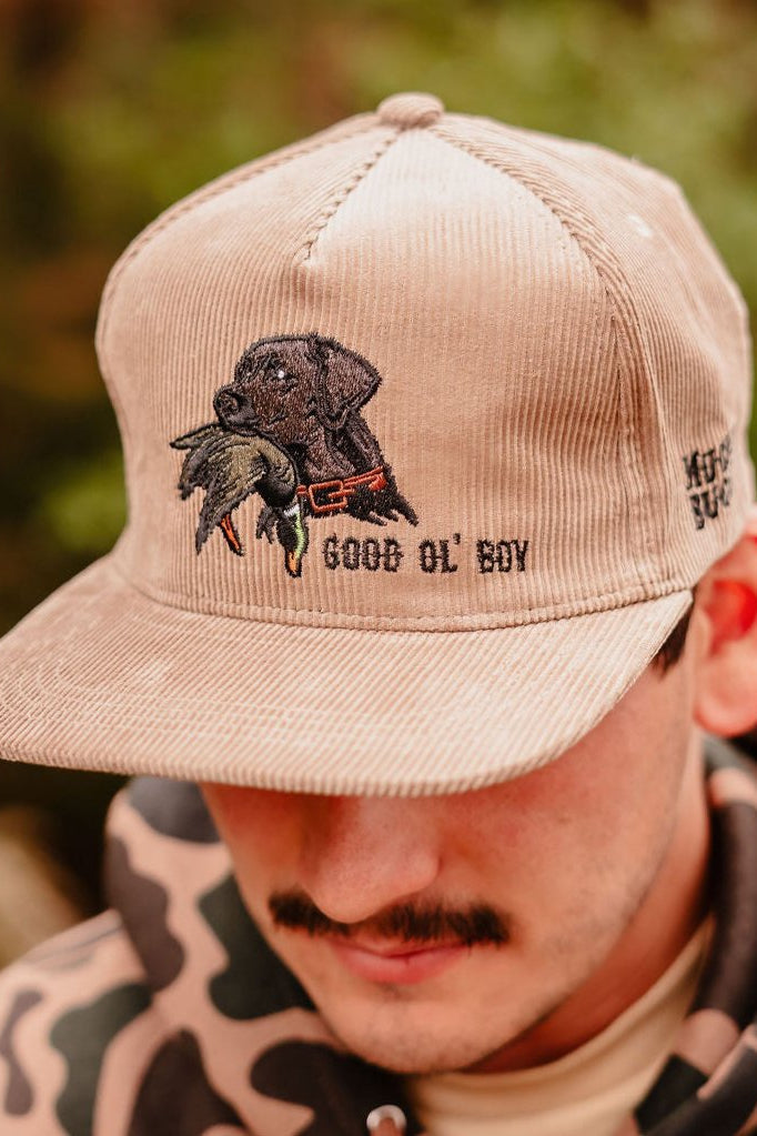 A person with a mustache and camo jacket wears the Mucker Bucket Good Ol' Boy Tan Corduroy snapback, featuring an embroidered black lab holding a duck and the words GOOD OL BOY on the front.