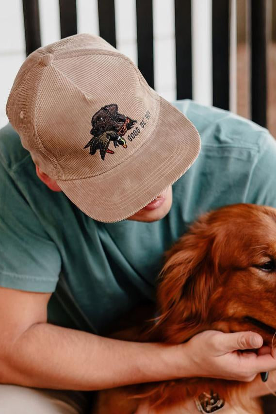 Person wearing a cap with a dog graphic, sitting outdoors with a brown dog.