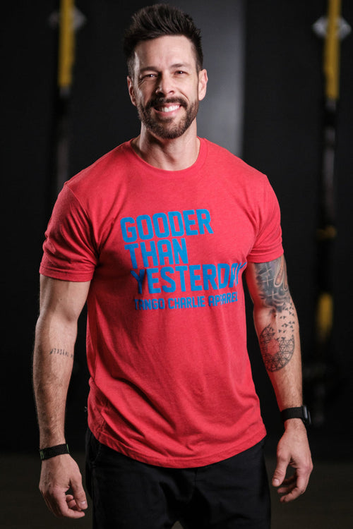 A bearded, tattooed man smiles in a gym wearing a Tango Charlie Apparel Gooder Than Yesterday red T-shirt and comfy AF black shorts. The soft polyester cotton blend of his outfit complements the blurred gym equipment in the background.