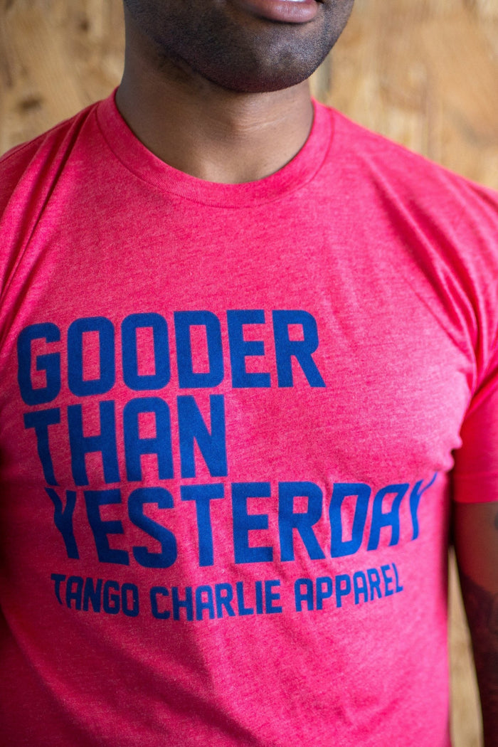 A person wears the Gooder Than Yesterday T-shirt by Tango Charlie Apparel, featuring blue text on a red background. Crafted from a comfy polyester-cotton blend, the vibrant design stands out against a wooden wall backdrop.