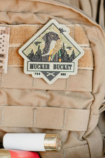 A Got One PVC Patch by Mucker Bucket, showing a dog with a stick, is attached to a tan tactical vest. Shotgun shells are lined along the bottom of the image.