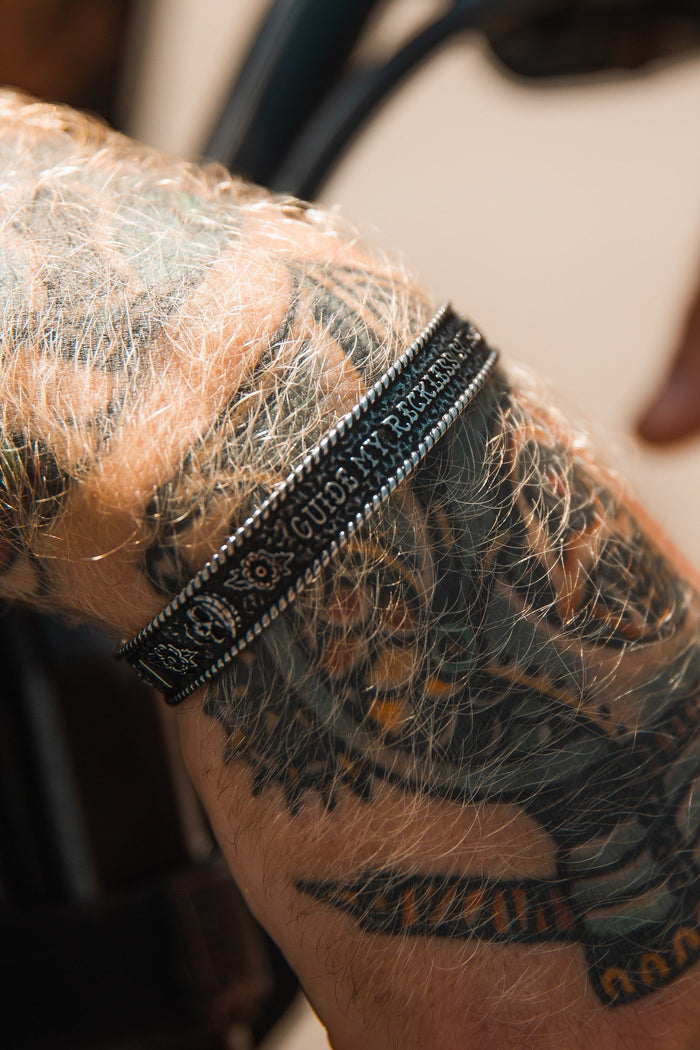 Close-up of a forearm with intricate bird head tattoos, adorned with the Guide My Reckless Soul Bracelet by Relentless Betrayal. This handcrafted black bracelet with detailed patterns stands out against light brown, sunlit, hairy skin, highlighting both the tattoo and bracelet artistry.